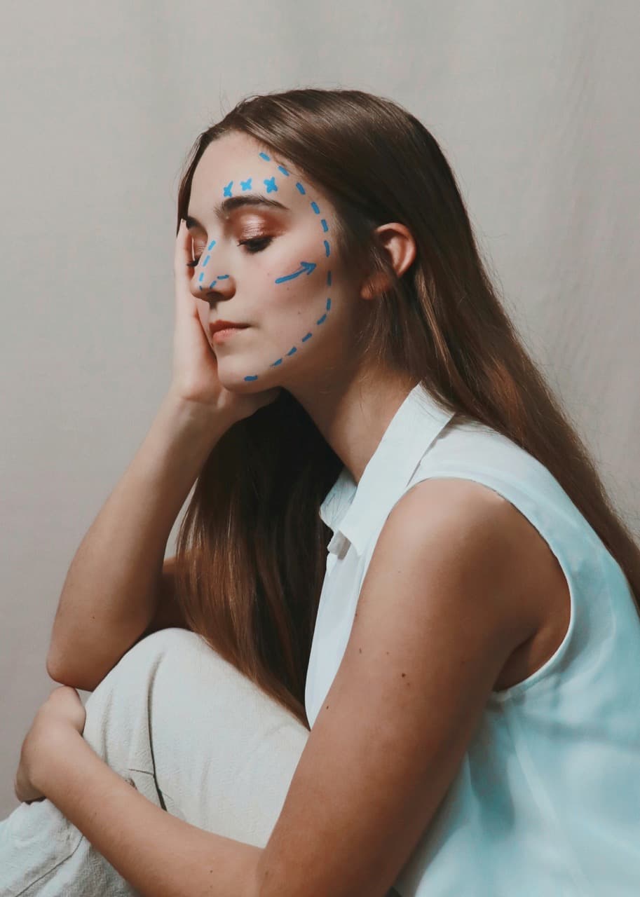 A woman with surgical markings on her face sits in contemplation, symbolizing cosmetic surgery planning. The image highlights the difference between wanting to “look better” and targeting specific facial features, reflecting expert approaches to aesthetic goals.