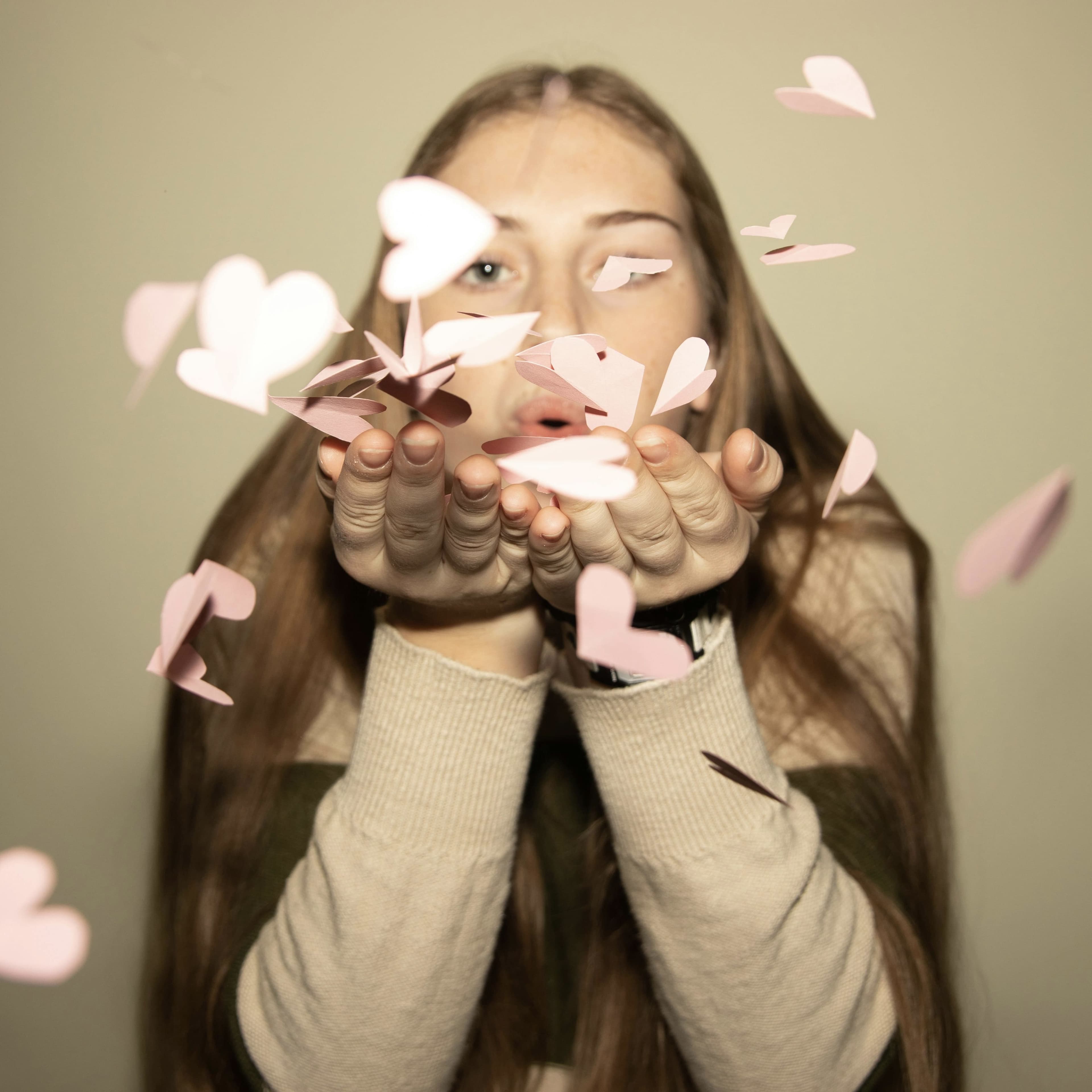 Young woman with long hair blowing pink paper hearts toward the camera, symbolizing love and celebration for Valentine’s Day.
